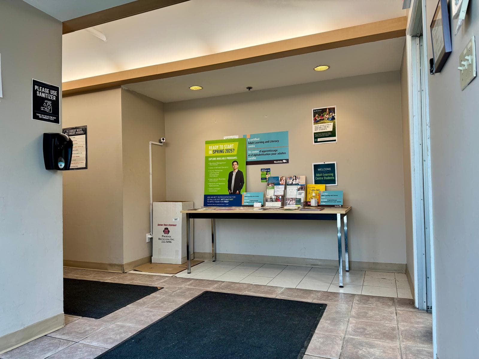 An entryway with dark floor mats, a table holding a standing sign and printed information sheets, and a wall-mounted sanitizer dispenser