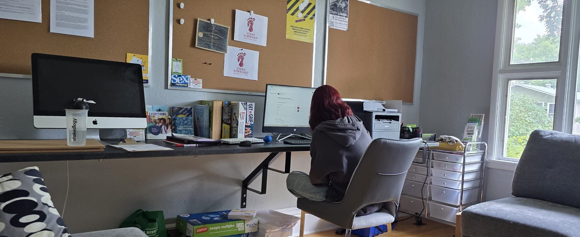 An office with two desktop computers on a long desk, a person working at one of them with their back to the camera, printed notices pinned to a board on the wall, and a window looking onto trees